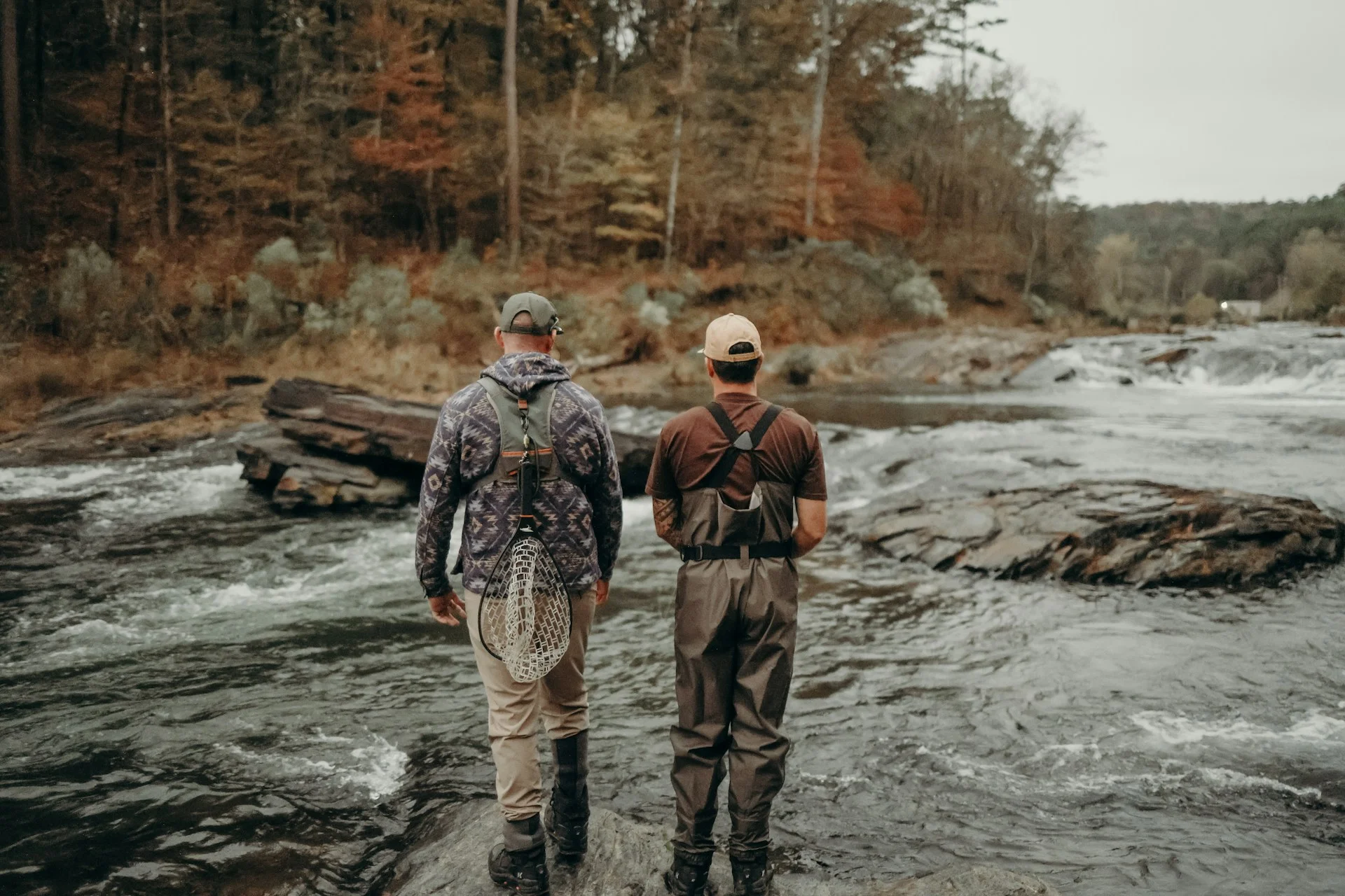 Two anglers in waders standing on rocks at the edge of an Oklahoma river, looking out over rapids with an autumn-colored hardwood forest behind them — representing the outdoor character and care for water in the Luther area